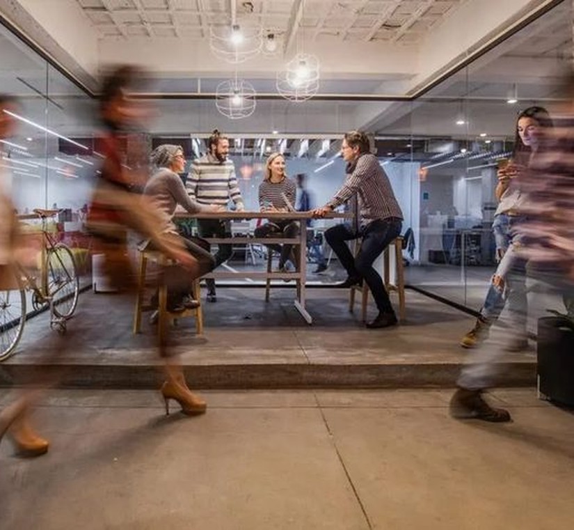 Personas conversando en un espacio de oficina con paredes de vidrio, con movimiento de gente al fondo